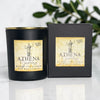 A matte black soy wax candle jar with a gold rim and a beige label reading 'Athena Goddess Burnt Fig Cassis' stands next to its matching black box on a light marble surface with blurred green leaves in the background.