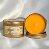 A round brass candle tin labeled BRIGID Goddess Sugar Magnolia, shown closed on the left and open on the right to display the bright orange wax, set against a draped white satin background.