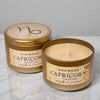 Two 'Oakmoss Capricorn' candles in decorative tins on a marble surface.