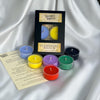 A boxed set of tea light candles from Victoria's Gardens, labeled 'Elemental Magick,' displayed on draped white satin with several small round candles in blue, yellow, purple, green, red, and black arranged around the package and an instruction card.