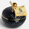 A black textured glass jar candle with a matching lid, accented by a gold band and a gold finial on the lid's handle. A paper tag from Victoria's Golds reads 'HEKATE Woodsy Soy Wax Candle' and is tied to the lid with twine.