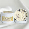 Two white metal tins of Kuan Yin Goddess Lotus Vanilla soy wax candle on a draped white fabric background. One tin is closed with a decorative label, and the other is open, showing the creamy off-white wax filled with dried botanical elements and a clear crystal.