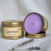 Lavender essential oil scented soy wax candle in a round gold metal tin, shown both closed and open on a white fabric background with lavender sprigs in front.