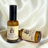 Two amber glass bottles of Mary Magdalene Natural Spray with black caps, one standing upright and the other lying on its side, set against a draped white satin fabric background.