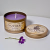 Scented candle in a metal tin labeled 'Mother Goddess' on a white surface with a purple flower.