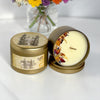 Two gold metal tins of 'Secret Garden' vanilla scented candles on a white surface, with one tin open to show the cream-colored wax and a colorful floral pattern on the inner lid.