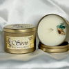 A gold metal tin candle shown open on draped white fabric, with the lid beside it. The candle wax is creamy off-white, and the lid label reads 'Siren' with 'Sea Glass' and 'Terra Nova' visible.