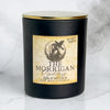 A matte black soy wax candle jar with a gold lid and a textured beige label reading 'The Morrigan Goddess Oakmoss Soy Wax Candle' by Victoria's Goddesses, set on a white marble surface.