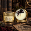Two gold tin candles displayed on a rustic wooden surface with gothic-themed props. One candle is closed, showing a cream-colored wax label with 'OAKMOSS THE MORRIGAN' and 'SOYWAX CANDLE' printed on it.