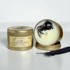 A soy wax candle in a round gold metal tin, shown closed with a label reading 'Oakmoss The Morrigan Fantasy' and next to an open tin displaying the cream-colored wax with a black spider design inside. A dark feather rests in front on a white surface against a light gray background.