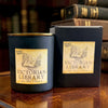 A dark navy blue soy wax candle jar with a gold rim and a vintage-style cream label reading 'Victorian Library' sits on a wooden table next to its matching dark box. The background shows a blurred library setting with old books.